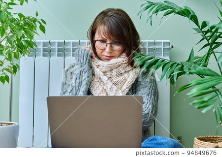 Woman in warm sweater using laptop sitting near heating radiator at home Woman in warm sweater using laptop sitting near heating radiator at home 94849676