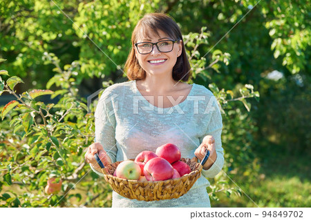 Smiling woman with harvest of ripe red apples in basket, outdoor garden 94849702