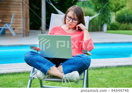 Middle aged woman sitting in chair in backyard with laptop 94849746