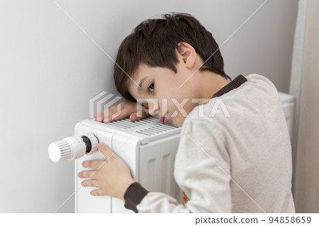 The child hugs the radiator of the home heater with his hands to keep warm. 94858659