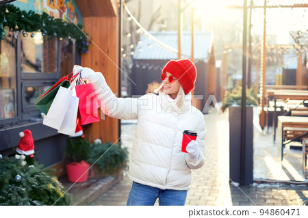 Woman walks around the city with a bunch of gift bags and drinks coffee from a disposable cup from a urban cafe. Coffee break at food kiosks after heavy shopping. choosing gifts for xmas and new year Woman walks around the city with a bunch of gift bags and drinks coffee from a disposable cup from a urban cafe. Coffee break at food kiosks after heavy shopping. choosing gifts for xmas and new year 94860471