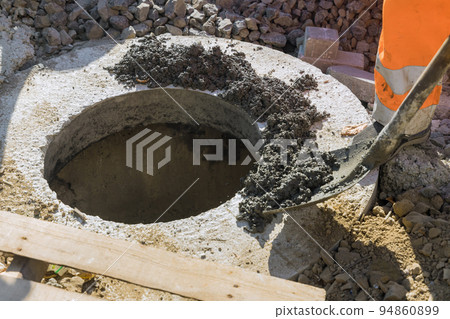 At a construction site, a cast-iron sewer hatch is being installed on a concrete base as part of the installation of a water sewer well in the ground to collect waste water 94860899