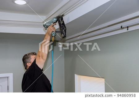 An experienced carpenter is seen in nailing wooden crown molding between the ceiling in a newly constructed dwelling An experienced carpenter is seen in nailing wooden crown molding between the ceiling in a newly constructed dwelling 94860958