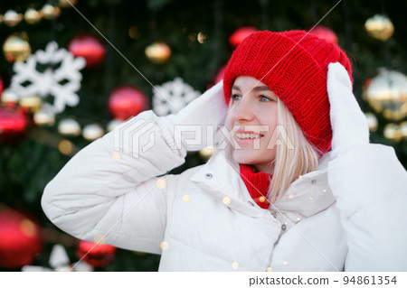 Beautiful blonde woman in a red knitted hat and a white jacket with mittens stands against the background of a Christmas tree in the city. Girl looks aside copy space. Celebrating New Year and xmas Beautiful blonde woman in a red knitted hat and a white jacket with mittens stands against the background of a Christmas tree in the city. Girl looks aside copy space. Celebrating New Year and xmas 94861354
