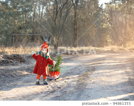 Small Christmas tree from the forest in its toy car is being driven by a kid along the path on a frosty morning on a sunny winter day.Retro clothes for a girl with a fur hat.Preparing for the New year Small Christmas tree from the forest in its toy car is being driven by a kid along the path on a frosty morning on a sunny winter day.Retro clothes for a girl with a fur hat.Preparing for the New year 94861355