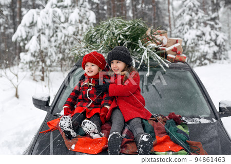 Cheerful children are sitting on the hood of a black car, on the roof of which there is a Christmas tree from the market and gifts with a red ribbon. Preparation for the celebration of New Year. Cheerful children are sitting on the hood of a black car, on the roof of which there is a Christmas tree from the market and gifts with a red ribbon. Preparation for the celebration of New Year. 94861463