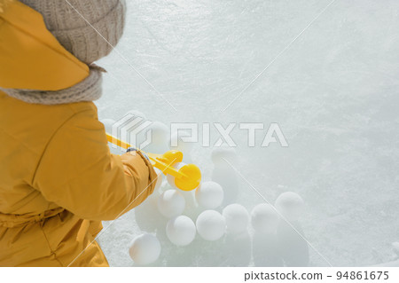A child makes snowballs out of snow with the help of a toy-a tool for making snowballs. PEOPLE FROM BEHIND. Winter frosty sunny day in nature. The girl holds the tongs in her hands. 94861675
