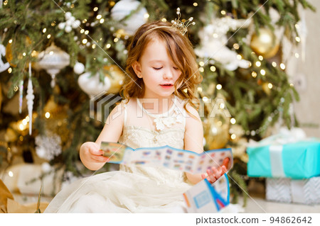 little girl reads the instruction from the box of gift by Christmas tree. little girl reads the instruction from the box of gift by Christmas tree. 94862642