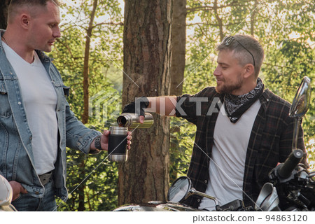Two man friends of motorcyclists bikers on motorcycles drinking tea from a thermos on the side of the road and talking Two man friends of motorcyclists bikers on motorcycles drinking tea from a thermos on the side of the road and talking 94863120
