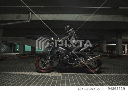 Young woman biker in a helmet sitting on a motorcycle in the underground parking garage Young woman biker in a helmet sitting on a motorcycle in the underground parking garage 94863319