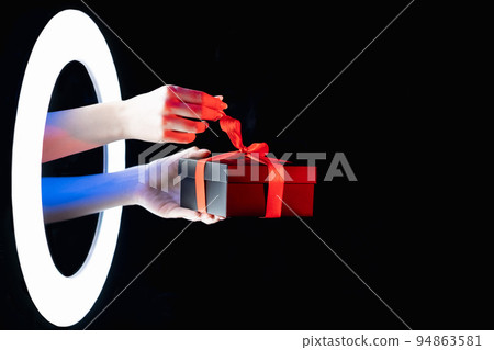Holiday surprise. Black Friday sale. Giveaway promotion. Female hands untying gift box with red ribbon in LED ring color neon light isolated on dark night empty space background. Holiday surprise. Black Friday sale. Giveaway promotion. Female hands untying gift box with red ribbon in LED ring color neon light isolated on dark night empty space background. 94863581