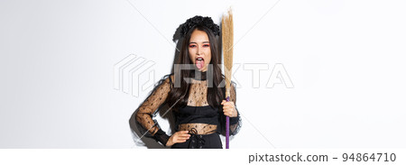 Carefree sassy asian woman enjoying halloween party, wearing witch costume and holding broom, showing tongue, standing over white background Carefree sassy asian woman enjoying halloween party, wearing witch costume and holding broom, showing tongue, standing over white background 94864710