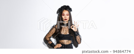 Sassy beautiful asian woman in witch costume showing okay gesture, all under control. Confident girl in halloween gothic dress like or approve something, standing white background 94864774