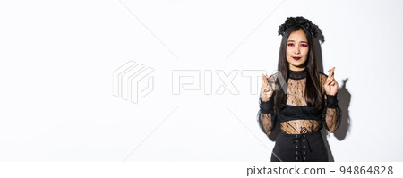 Image of hopeful asian girl in gothic lace dress and wreath making wish, cross fingers for good luck, looking wishful at camera, standing over white background 94864828