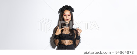 Image of curious beautiful asian girl in witch costume pointing finger at credit card, standing over white background 94864878