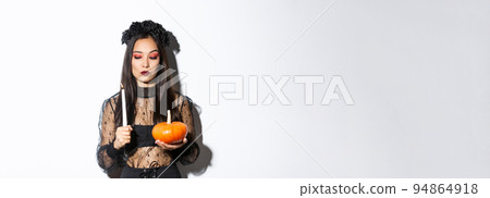 Myserious asian wicked witch in gothic dress, looking at lit candle, holding pumpkin, standing over white background 94864918