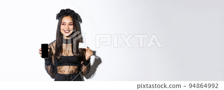 Dreamy smiling girl looking away while thinking, showing credit card and mobile phone, wearing halloween gothic dress Dreamy smiling girl looking away while thinking, showing credit card and mobile phone, wearing halloween gothic dress 94864992
