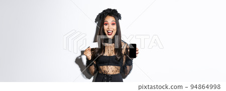 Cheerful beautiful asian woman in halloween costume, dressed as witch and showing smartphone with credit card, standing over white background 94864998