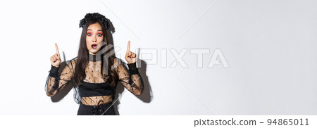 Impressed asian girl in black lace dress and wreat turn your attention on halloween promo, making announcement, pointing fingers up, showing banner, standing over white background Impressed asian girl in black lace dress and wreat turn your attention on halloween promo, making announcement, pointing fingers up, showing banner, standing over white background 94865011