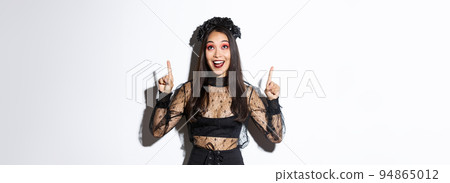 Image of amused and happy asian female magician, girl in witch costume pointing fingers at upper left corner, looking at your logo, celebrating halloween, standing over white background Image of amused and happy asian female magician, girl in witch costume pointing fingers at upper left corner, looking at your logo, celebrating halloween, standing over white background 94865012
