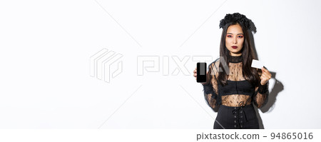 Serious-looking elegant young asian woman in gothic dress and black wreath showing credit card and mobile phone screen, celebrating halloween Serious-looking elegant young asian woman in gothic dress and black wreath showing credit card and mobile phone screen, celebrating halloween 94865016