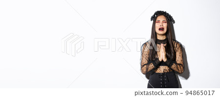 Miserable young asian woman in trouble pleading god, crying and begging for help, wearing halloween gothic dress and wreath, supplicating over white background 94865017