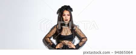 Angry and pissed-off asian female magician, evil witch in black dress and wreath looking mad at someone, frowning and looking judgemental, standing over white background 94865064