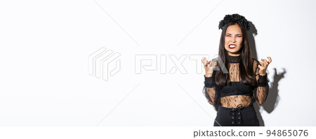 Image of angry wicked witch cursing and swearing, clenching fists and grimacing with hatred, standing over white background, celebrating halloween 94865076