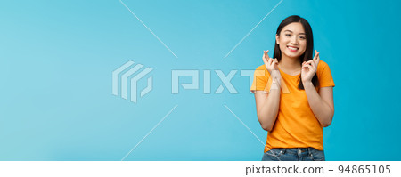 Cheerful smiling hopeful asian girl praying wish for good luck, cross fingers grinning toothy anticipate positive good news, stand blue background supplicating optimistic feelings hope win 94865105