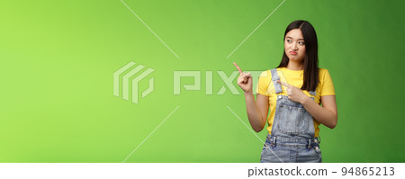 Upset cute asian female student pulling face regret shame, pity missing good chance buy product sale, pointing looking left disappointed, jealously gaze coworker, stand green background 94865213