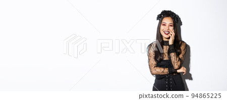 Image of happy good-looking asian woman in gothic dress and black wreath, laughing at halloween party, standing over white background Image of happy good-looking asian woman in gothic dress and black wreath, laughing at halloween party, standing over white background 94865225