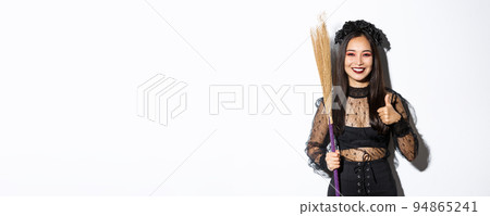 Image of smiling asian woman in witch costume with broom, showing thumbs-up in approval, standing over white background 94865241