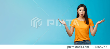 Questioned unaware young asian girl with dark short haircut shrugging hands spread sideways, look confused open mouth uncertain, being clueless, stand blue background wear yellow t-shirt 94865243