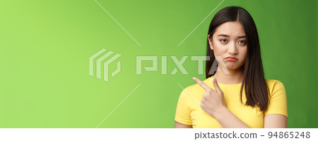 Grumpy upset cute asian girl complaining tired, drained during university exams, pointing unhappy, losing competition, pointing left, sulking displeased, stand green background reluctant 94865248
