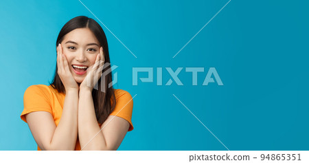 Flirty cute asian girl look surprised, express admiration and joy, touch cheeks, blushing coquettish smiling receive good news, stand blue background happily react amazed and excited 94865351