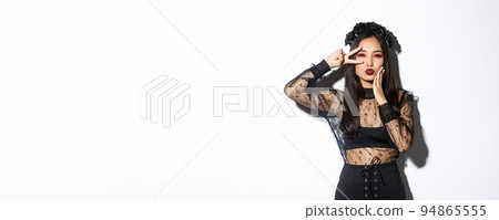 Image of beautiful asian woman in gothic lace dress and black wreath posing on halloween banner, showing peace gesture and pouting, standing over white background 94865555