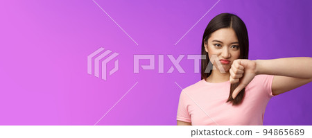 Close-up unsatisfied displeased judgemental asian girl give thumb down, grimacing, cringing from disappointment, not recommend awful product, complain bad service, stand purple background 94865689