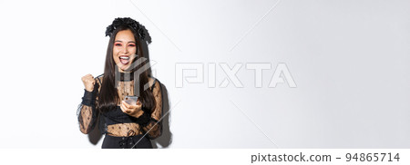 Image of successful happy asian woman celebrating good news, fist pump in rejoice and holding smartphone, wearing halloween costume, standing over white background 94865714