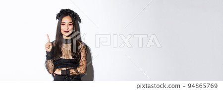 Creative young woman in witch costume smiling pleased as have great idea, raising finger to say suggestion. Female asian dressed-up like widow or mysterious magician, white background Creative young woman in witch costume smiling pleased as have great idea, raising finger to say suggestion. Female asian dressed-up like widow or mysterious magician, white background 94865769
