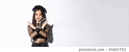 Confident beautiful woman in black gothic dress showing cross gesture, disapprove and stop something bad, disagree with someone about halloween, standing over white background 94865783