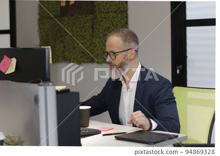 A European adult man with a beard works at a computer in the office of company A European adult man with a beard works at a computer in the office of company 94869328