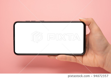 Hand holding mobile phone in horizontal position for mockup. watching streaming video on cellphone on pink table background. Close up of horizontal black smartphone with blank screen in woman hands Hand holding mobile phone in horizontal position for mockup. watching streaming video on cellphone on pink table background. Close up of horizontal black smartphone with blank screen in woman hands 94872357