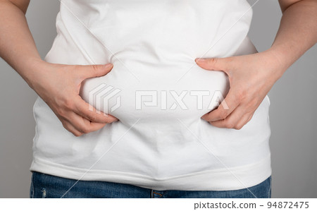 Woman with fat flabby belly, overweight female body on gray background. Woman in jeans and a light shirt is standing sideways and holding hands squeezes belly fat. Weight loss, diet lifestyle Woman with fat flabby belly, overweight female body on gray background. Woman in jeans and a light shirt is standing sideways and holding hands squeezes belly fat. Weight loss, diet lifestyle 94872475