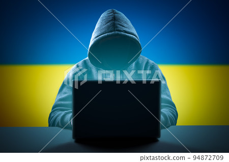 Faceless hooded anonymous computer hacker on Ukrainian flag background. Internet crime and electronic banking security. Anonymous person in the hood sitting in front of computer working with laptop 94872709
