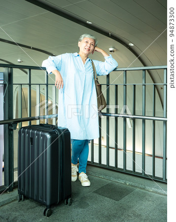 60多歲的亞洲熟年婦女微笑著站著,身旁有手提箱行李袋和手持登機證與護照,適用於covid19疫情後旅 94873300