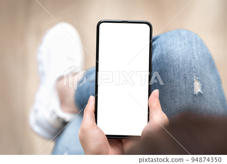 Top view mockup image of woman holding mobile phone with blank white screen. Woman in jeans sits cross-legged and holds cell phone in her hands with blank white screen 94874350