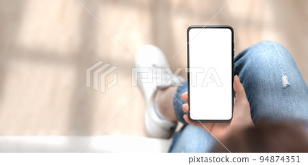Top view mockup image of woman holding mobile phone with blank white screen. woman using cellphone with while sitting cross-legged on sofa at home. Mockup smartphone. wide image banner 94874351