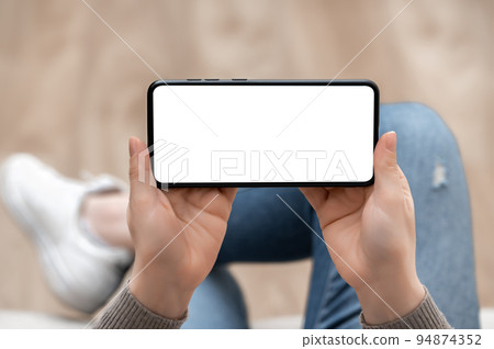Top view mockup image of woman holding mobile phone horizontally with blank white screen. woman using cellphone horizontal with while sitting cross-legged on sofa at home. Mockup smartphone 94874352