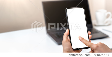 Mockup cellphone and laptop. Woman hand holding phone with blank screen in office. Woman finger presses on screen smartphone. wide banner Mockup cellphone and laptop. Woman hand holding phone with blank screen in office. Woman finger presses on screen smartphone. wide banner 94874387