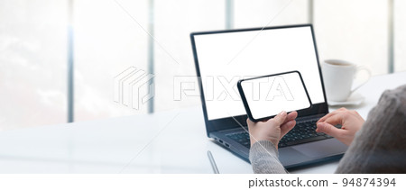 Mockup image of woman using laptop and phone with blank white screen on white table in modern loft cafe. hand holding smartphone horizontally with white screen in office 94874394